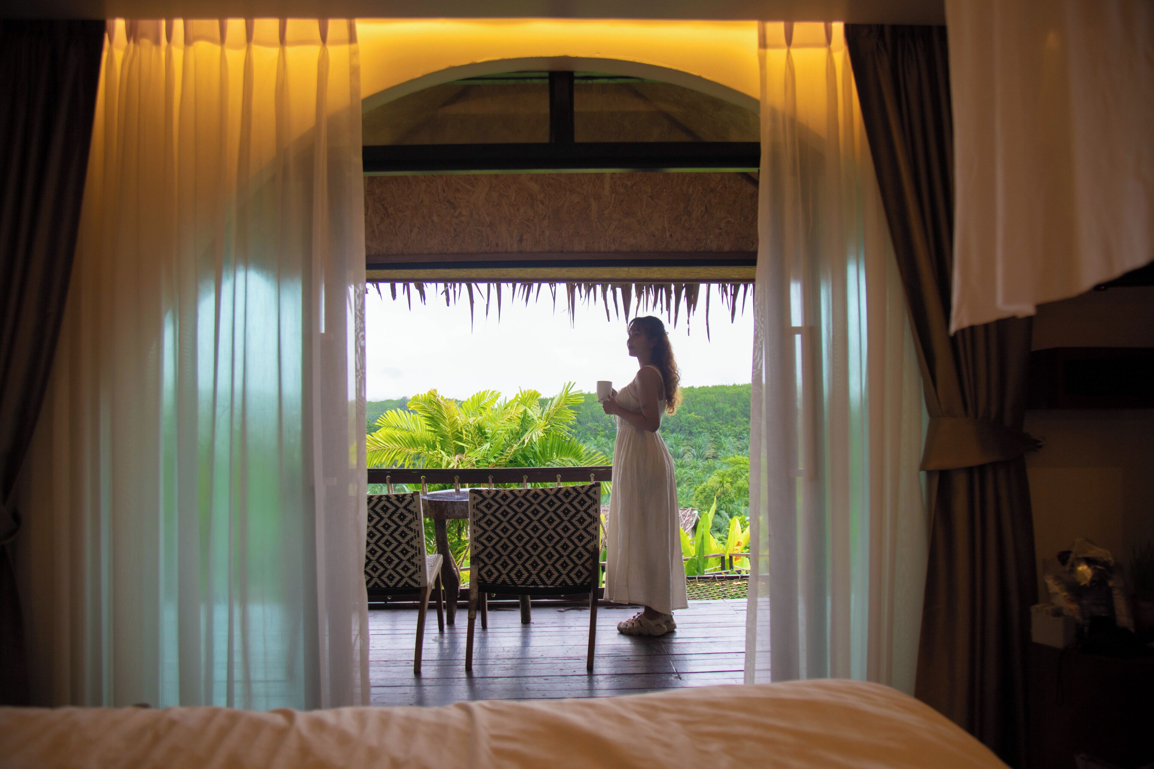 Una habitación con cama, una mesa y vistas a un paisaje verde y exuberante a través de una ventana con cortinas.