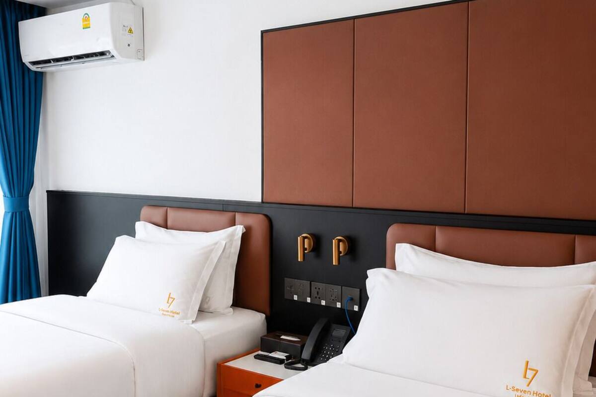 Una habitación de hotel con dos camas, un cabecero marrón, un aire acondicionado instalado en la pared y una mesita de noche con un teléfono y una lámpara.