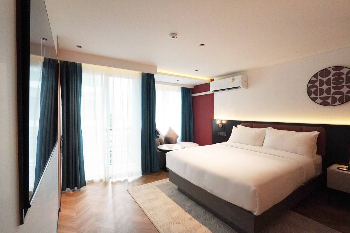 Una habitación de hotel moderna con una cama grande, una mesita de noche, un reloj de pared redondo y una ventana con cortinas.