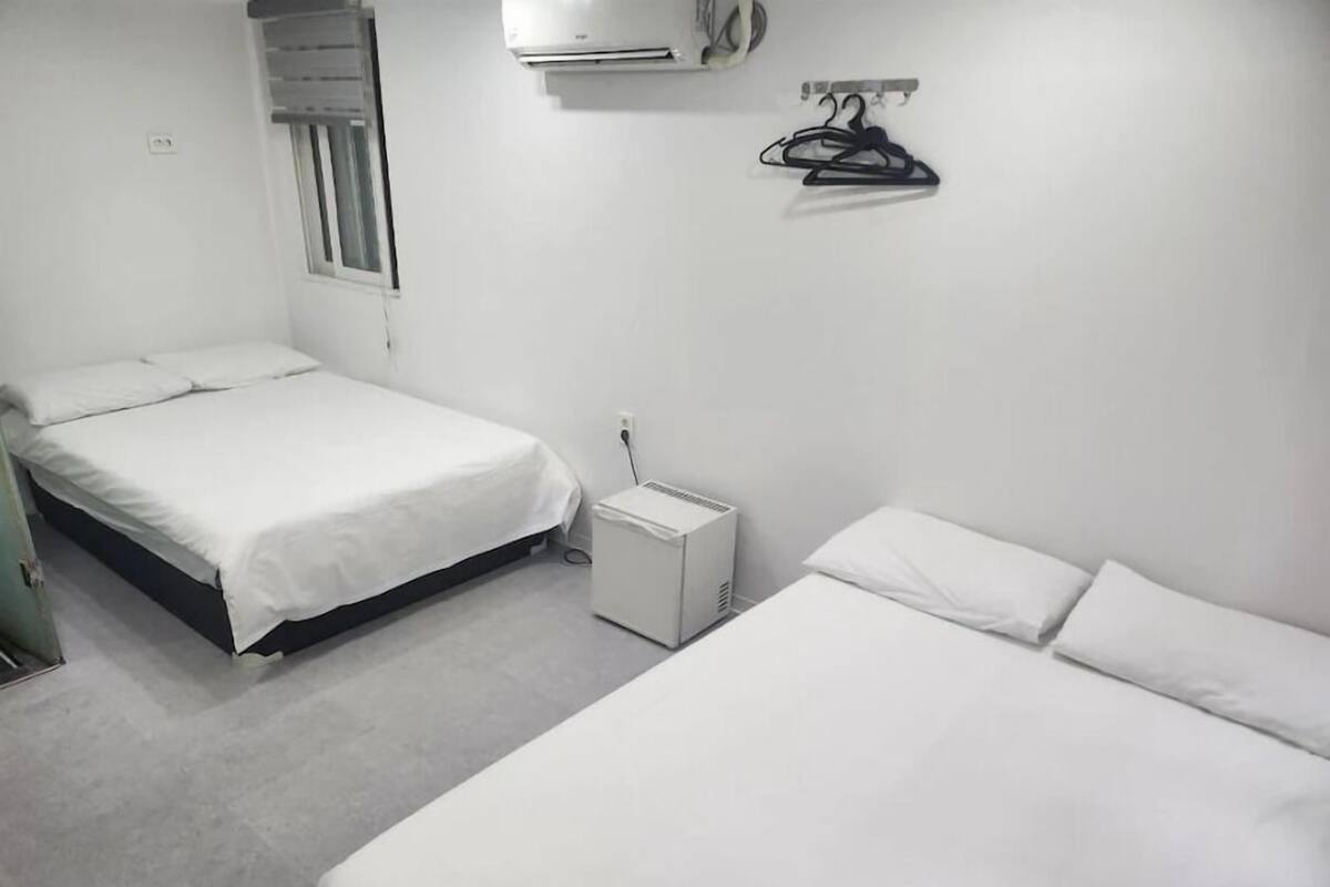 Una habitación con dos camas, una mesita de noche pequeña, un aire acondicionado, una ventana con persianas y un perchero en la pared.