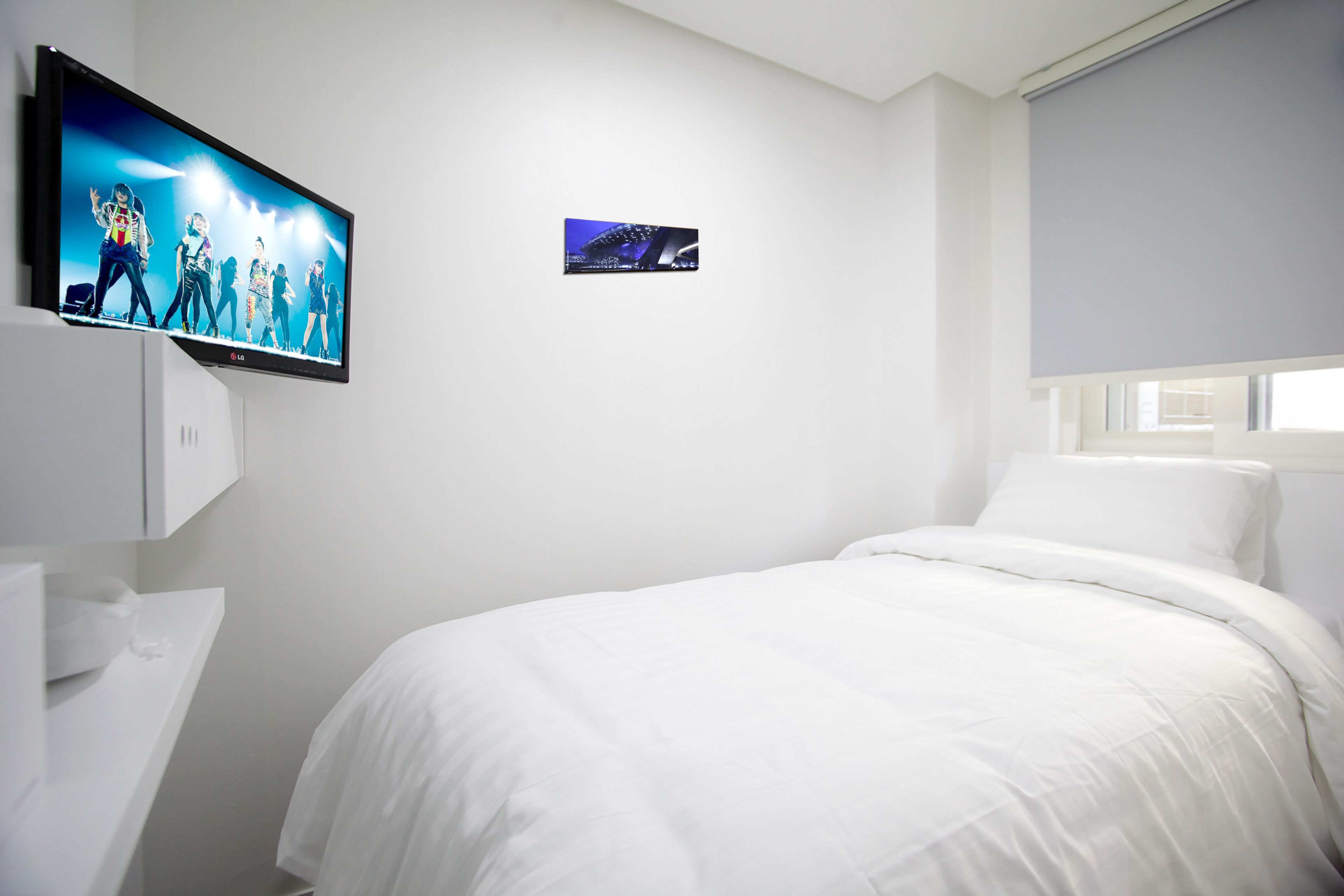 A hotel room with a large flat-screen TV mounted on the wall, a small picture frame, a white bed, and a window with a blind.