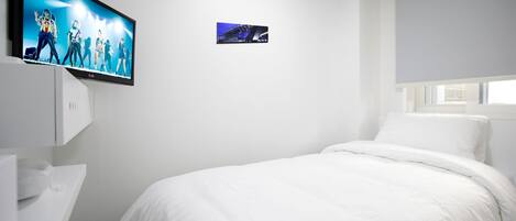 A hotel room with a large flat-screen TV mounted on the wall, a small picture frame, a white bed, and a window with a blind.