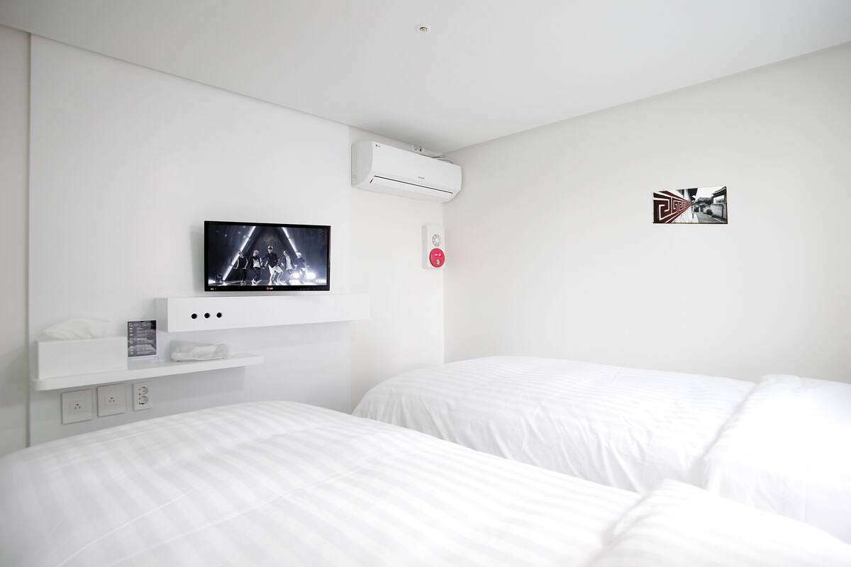 Una habitación de hotel moderna con televisión de pantalla plana en la pared, unidad de aire acondicionado integrada, mesita de noche con lámpara y cama con ropa blanca.