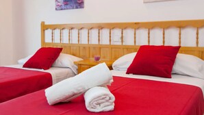 Ein Hotelzimmer mit zwei Betten, beide mit roter Bettwäsche und weißen Kissen. Auf den Betten liegen Handtücher und ein kleiner Tisch mit einem violetten Gegenstand darauf.