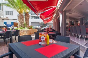 Un área de comedor al aire libre con mesas, sillas y una sombrilla roja, además de un atril para el menú.