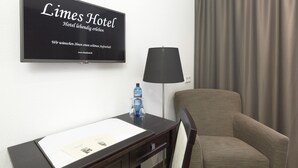 A hotel room with a flat-screen TV displaying the hotel's name and website, a desk with a chair, a lamp, and a bottle of water. - Limes Hotel (Wehrheim)