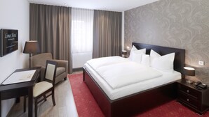 A hotel room with a large bed, a desk with a chair, a nightstand with a lamp, and a window with curtains. - Limes Hotel (Wehrheim)
