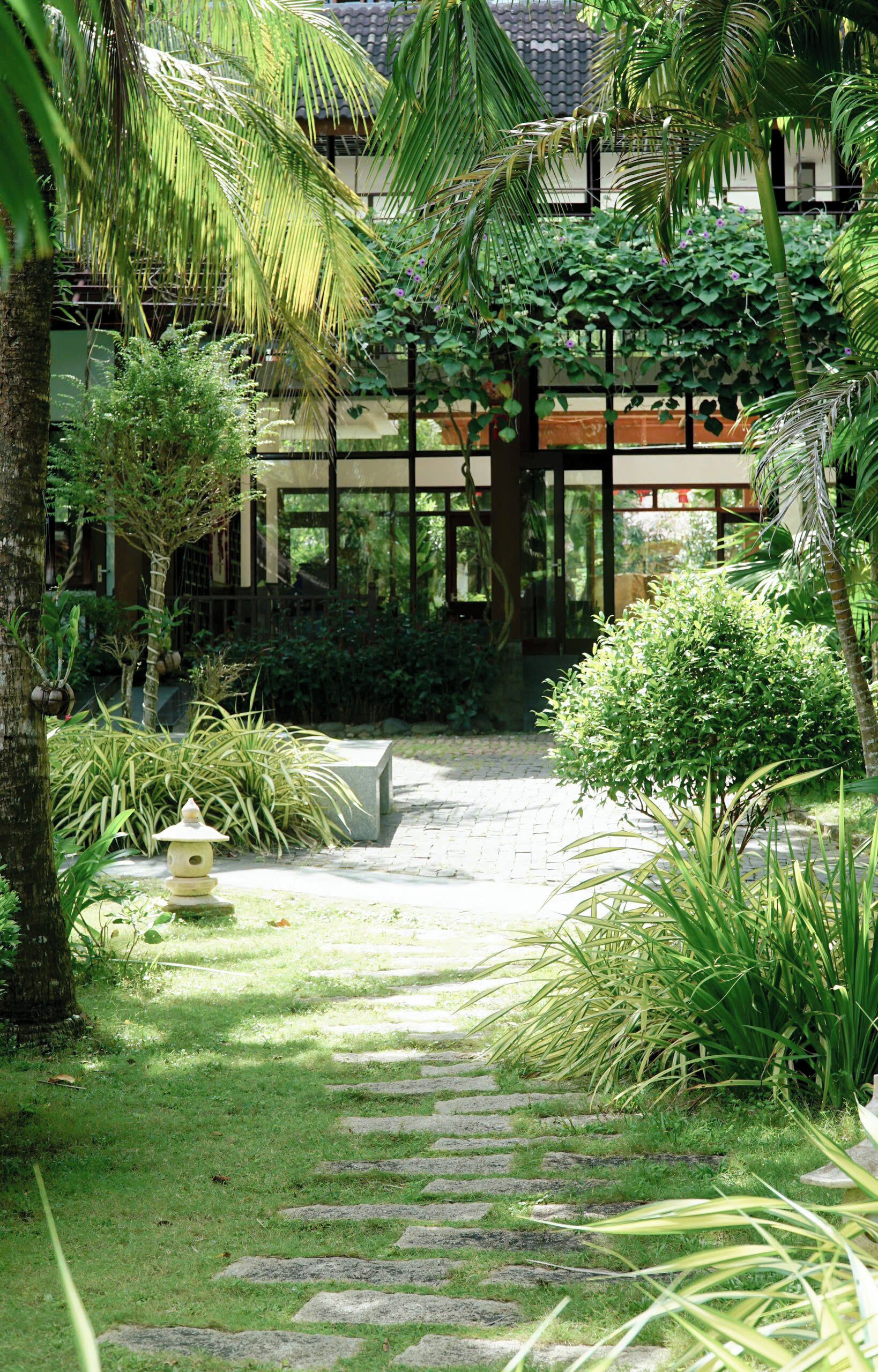 Un vialetto nel giardino conduce a un edificio con un tetto tradizionale, circondato da vegetazione rigogliosa e un farolino in pietra.