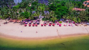 An aerial view of a beach with rows of sun loungers and umbrellas, surrounded by palm trees and buildings in the background. - Tropicana Resort (Phu Quoc)