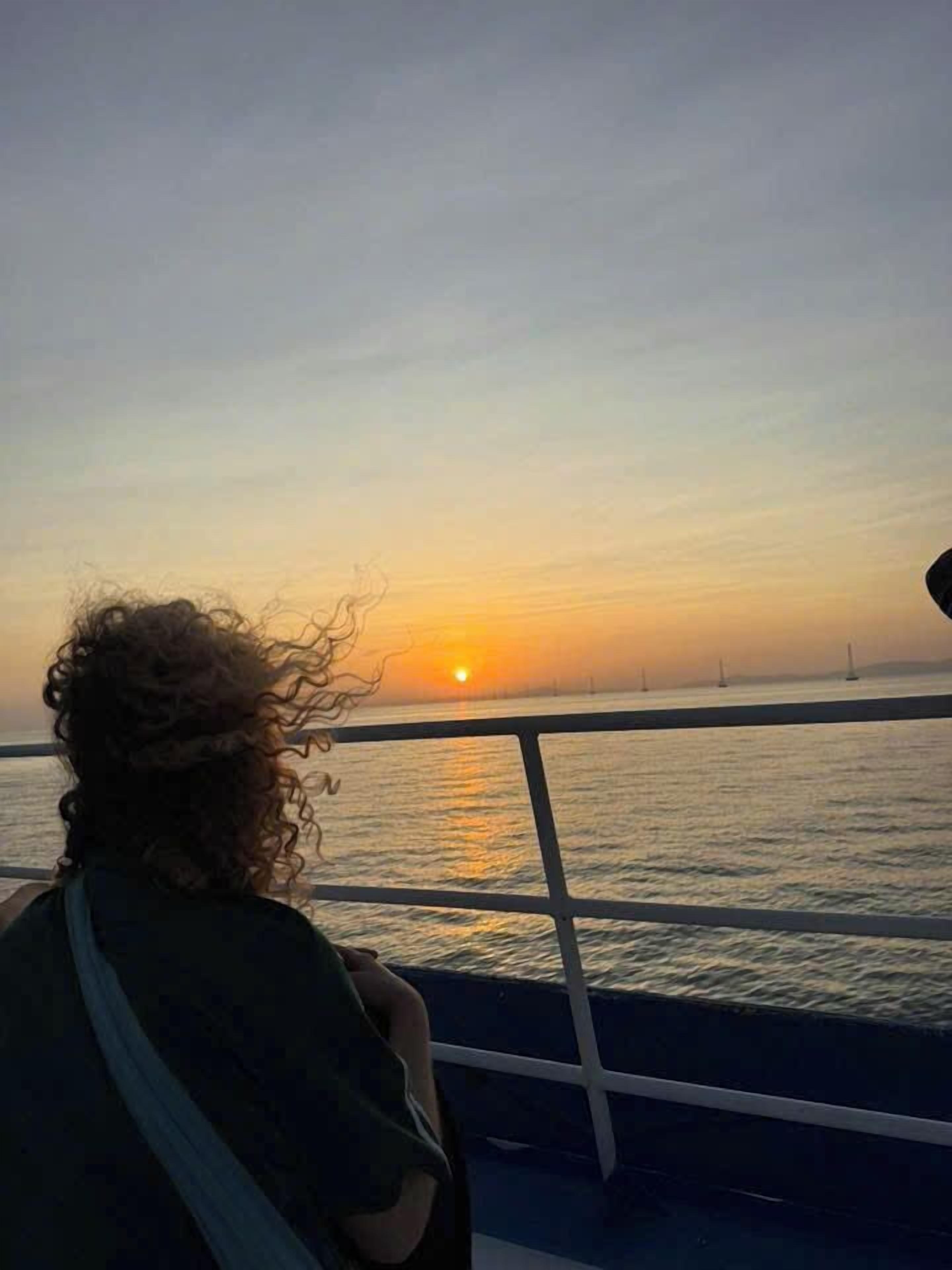 A person with curly hair sitting by a railing, overlooking a sunset over water with sailboats in the distance.