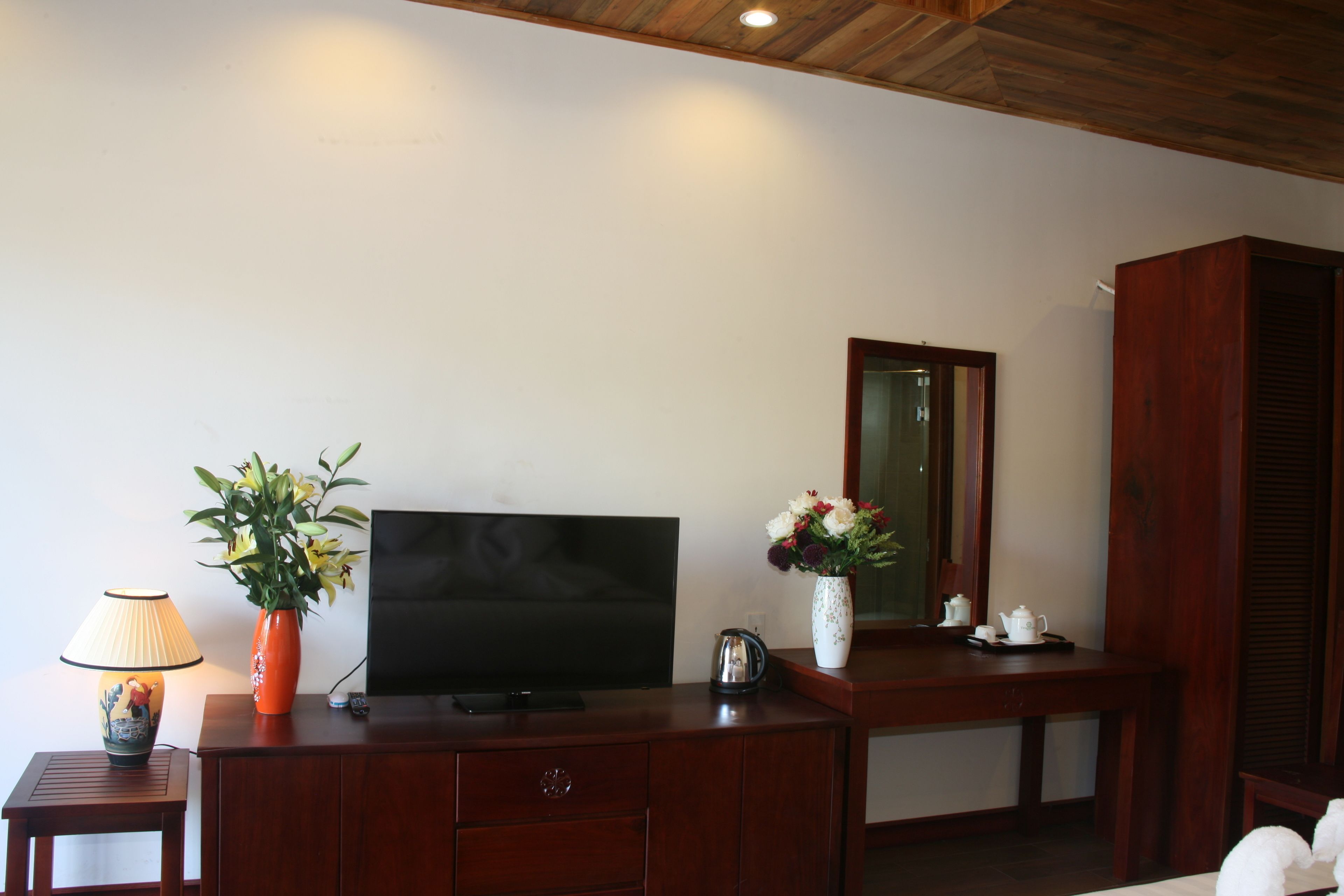 Une pièce avec un bureau en bois, une télévision, un vase de fleurs, une lampe et un miroir.