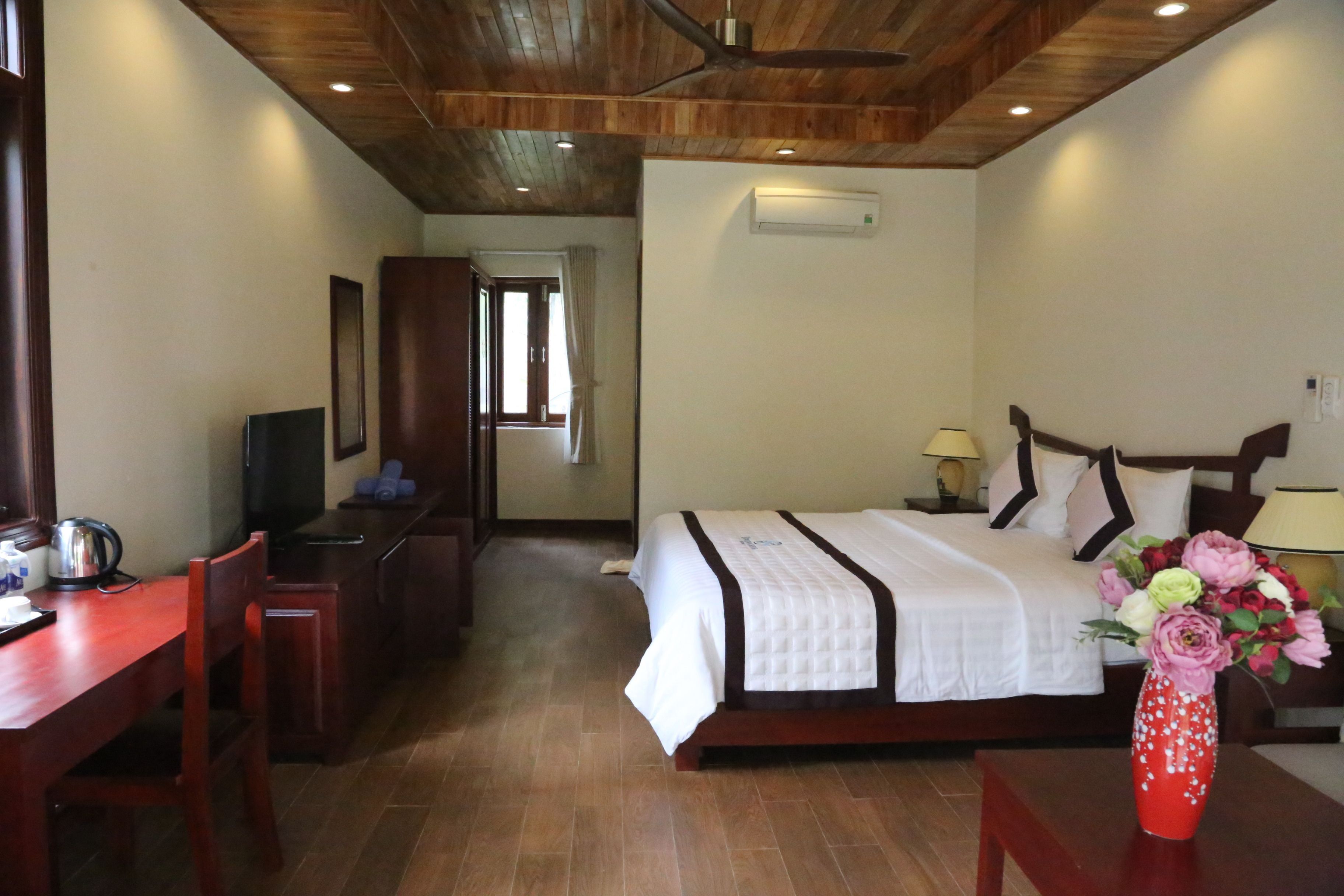 A hotel room with a wooden ceiling, a bed with white bedding, a desk with a chair, a TV, and a vase with flowers.