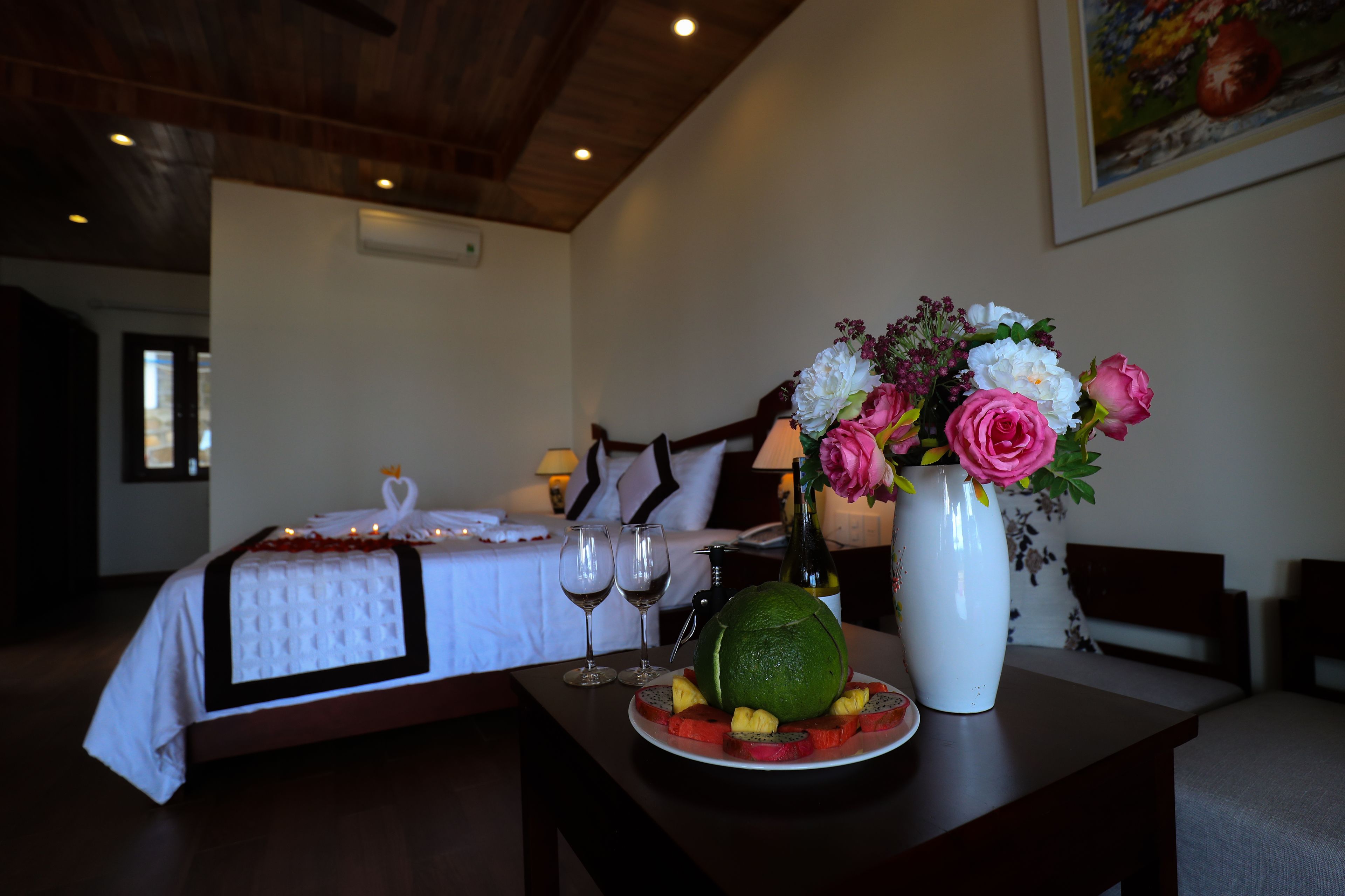 A hotel room with a neatly made bed, a dining table with a fruit platter and a vase of flowers, and a painting on the wall.