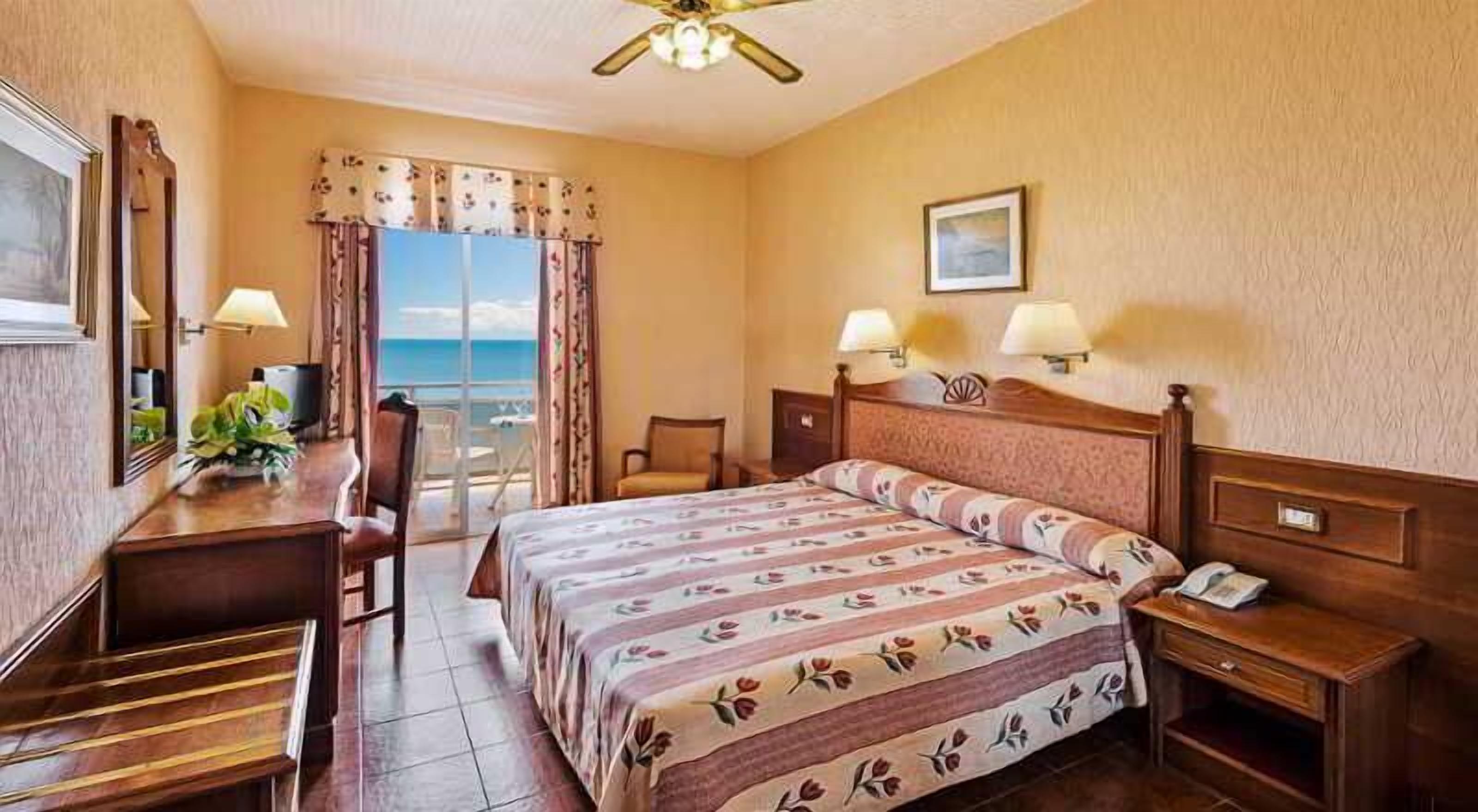Habitación de hotel con cama, escritorio, silla, mesita de noche, lámpara, ventilador de techo y ventana con vistas.