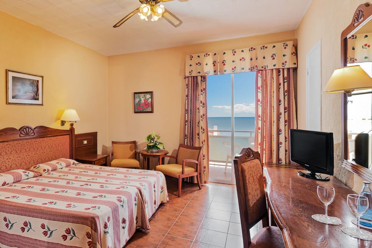 Ein Hotelzimmer mit einem Bett, zwei Stühlen, einem Schreibtisch mit Fernseher, einem kleinen Tisch mit Pflanze und einem Balkon mit Vorhängen.