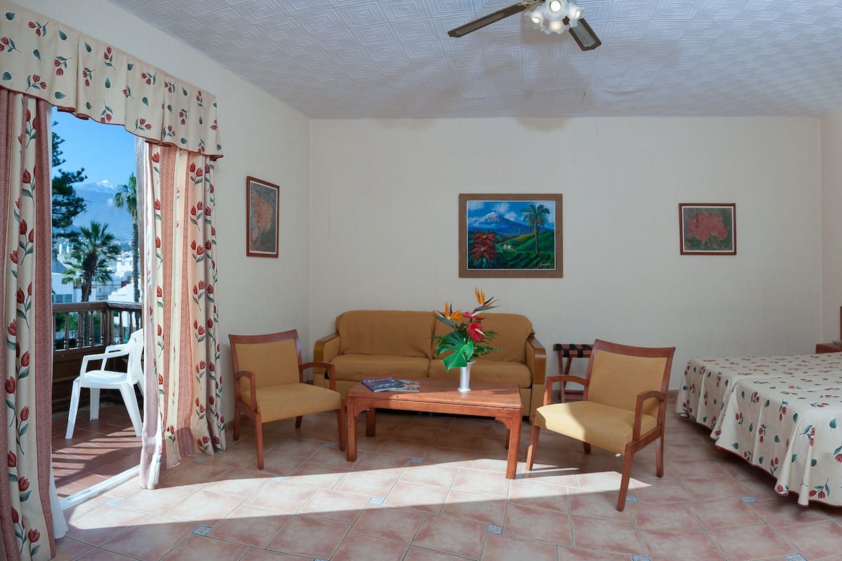 Ein Hotelzimmer mit einem Bett, zwei Stühlen, einer Couch, einem kleinen Tisch mit Blumenvase und einem Balkon mit Blick auf Palmen und ein Gebirge.