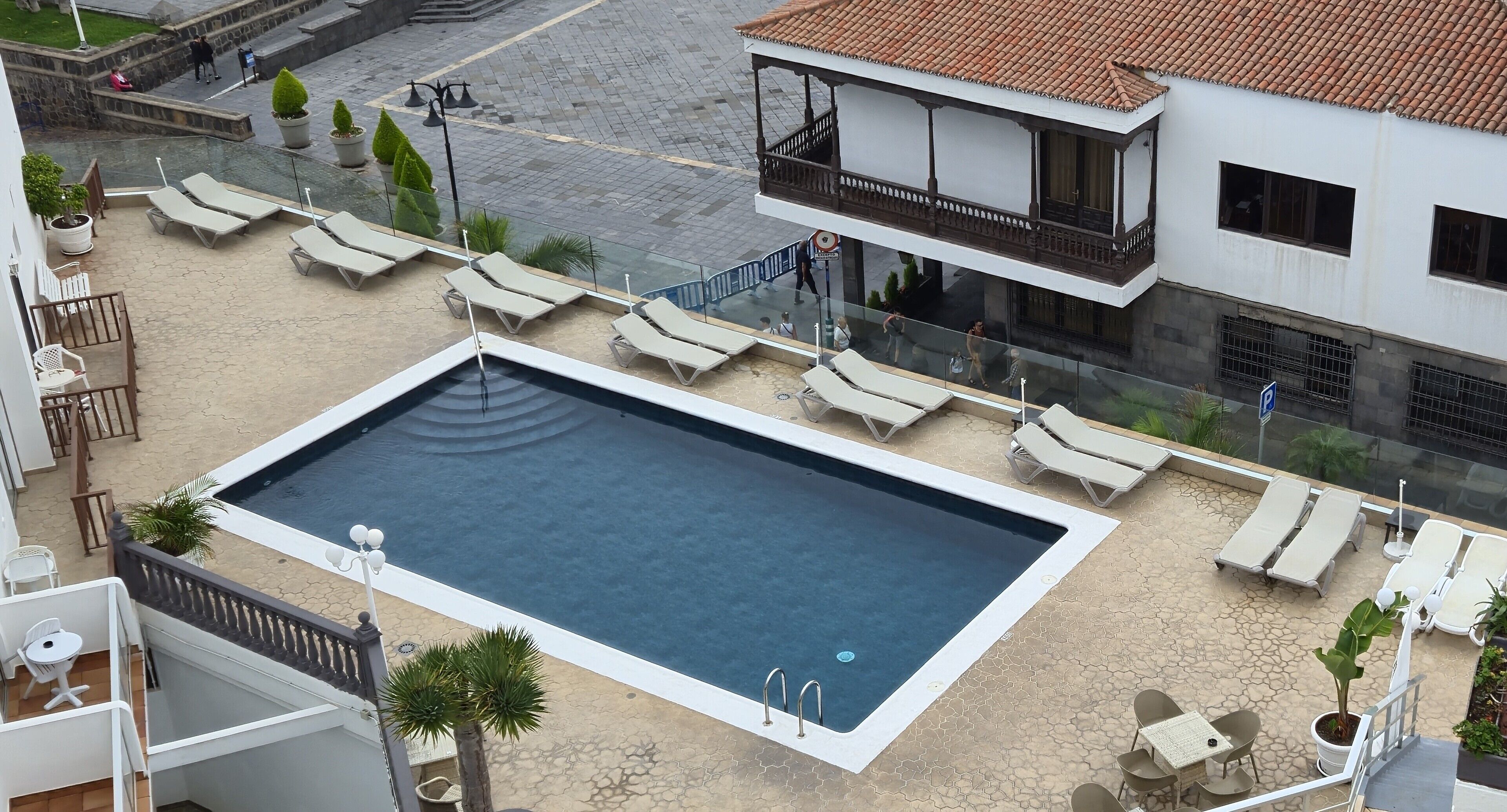 A rooftop swimming pool with lounge chairs and a small table, surrounded by potted plants and a building with a balcony.
