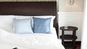 Una cama bien hecha con ropa blanca y dos almohadas azules, una mesita de noche con un vaso y una lámpara de pared.