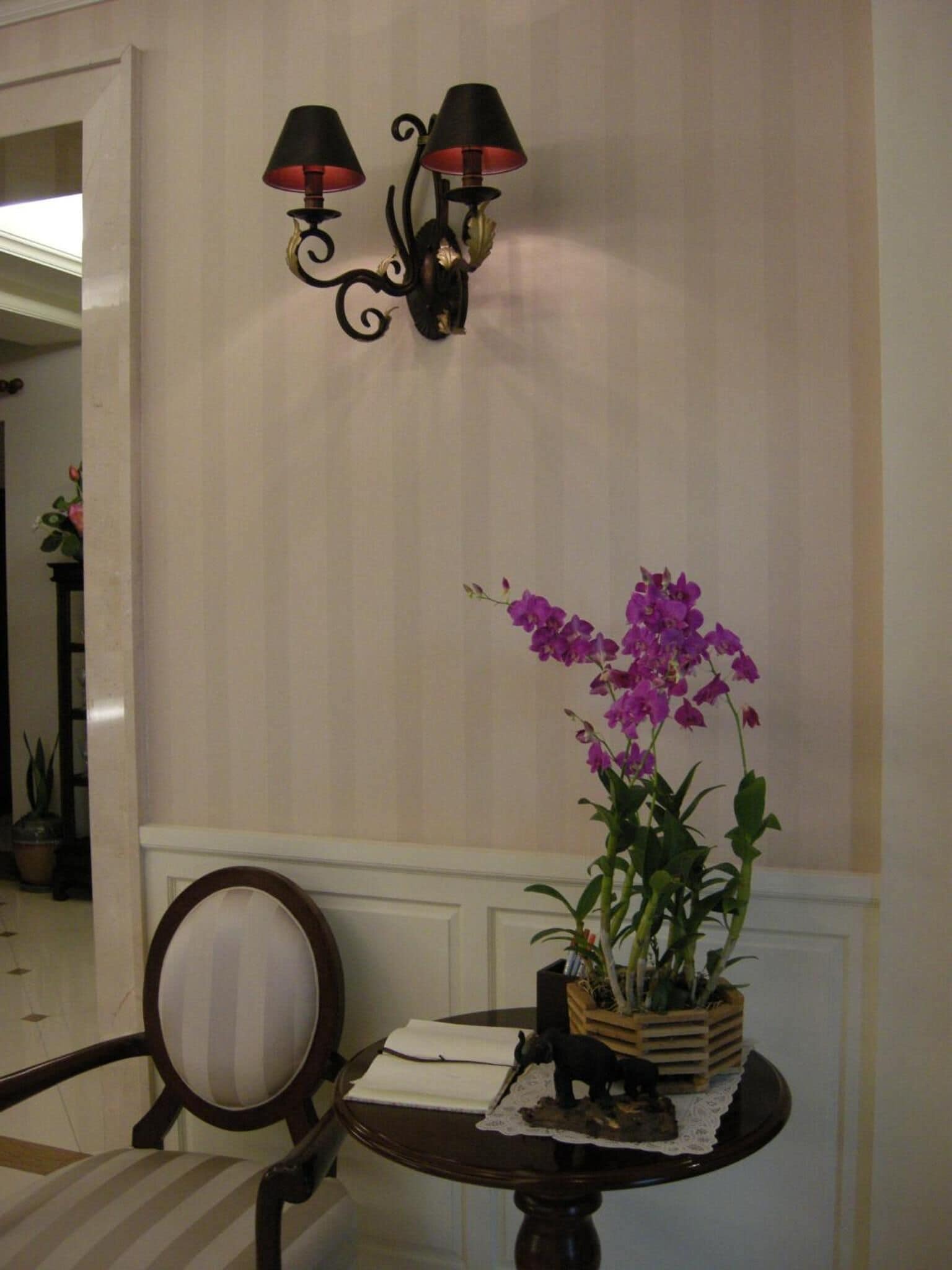A room with a table featuring a potted orchid, a small figurine, and a notebook. There are two wall-mounted lamps with black shades.