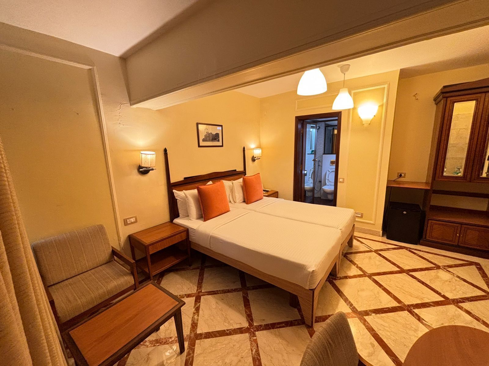Quarto de hotel com cama, mesas de cabeceira, uma cadeira, escrivaninha e banheiro visível por uma porta aberta, além de um espelho.