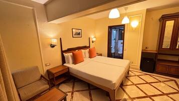 Quarto de hotel com cama, mesas de cabeceira, uma cadeira, escrivaninha e banheiro visível por uma porta aberta, além de um espelho.
