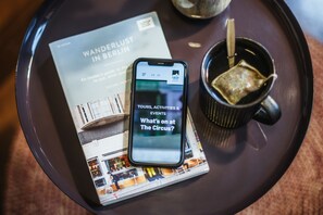 A smartphone displaying a travel app on a tray with a magazine and a cup. - The Circus Hotel (Berlin)
