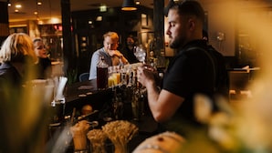 A bar area with a bartender preparing drinks, a counter with various bottles and glasses, and hanging pendant lights. - The Circus Hotel (Berlin)