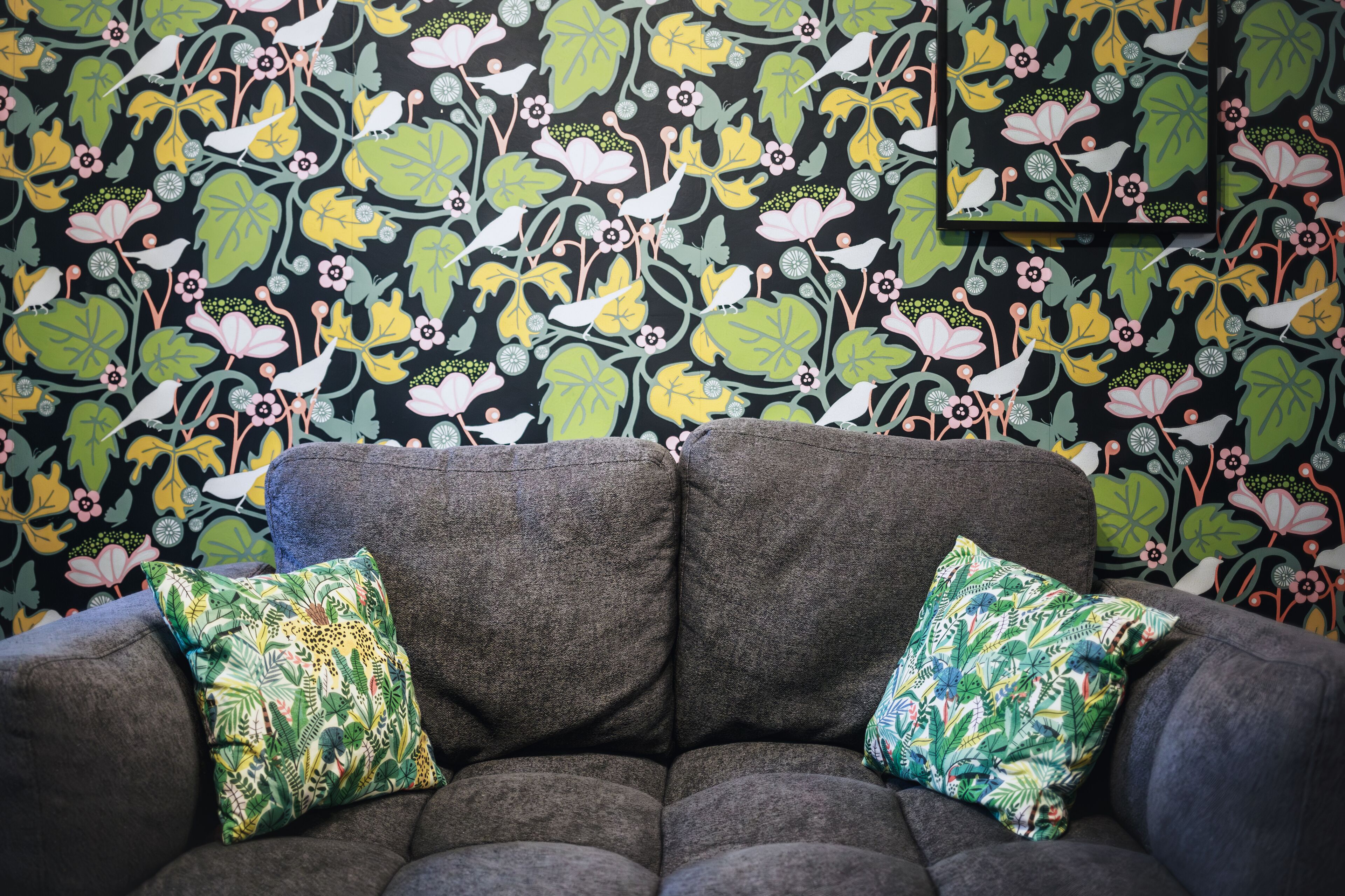 Un sofá gris con dos cojines estampados frente a una pared decorada con motivos florales y aves.