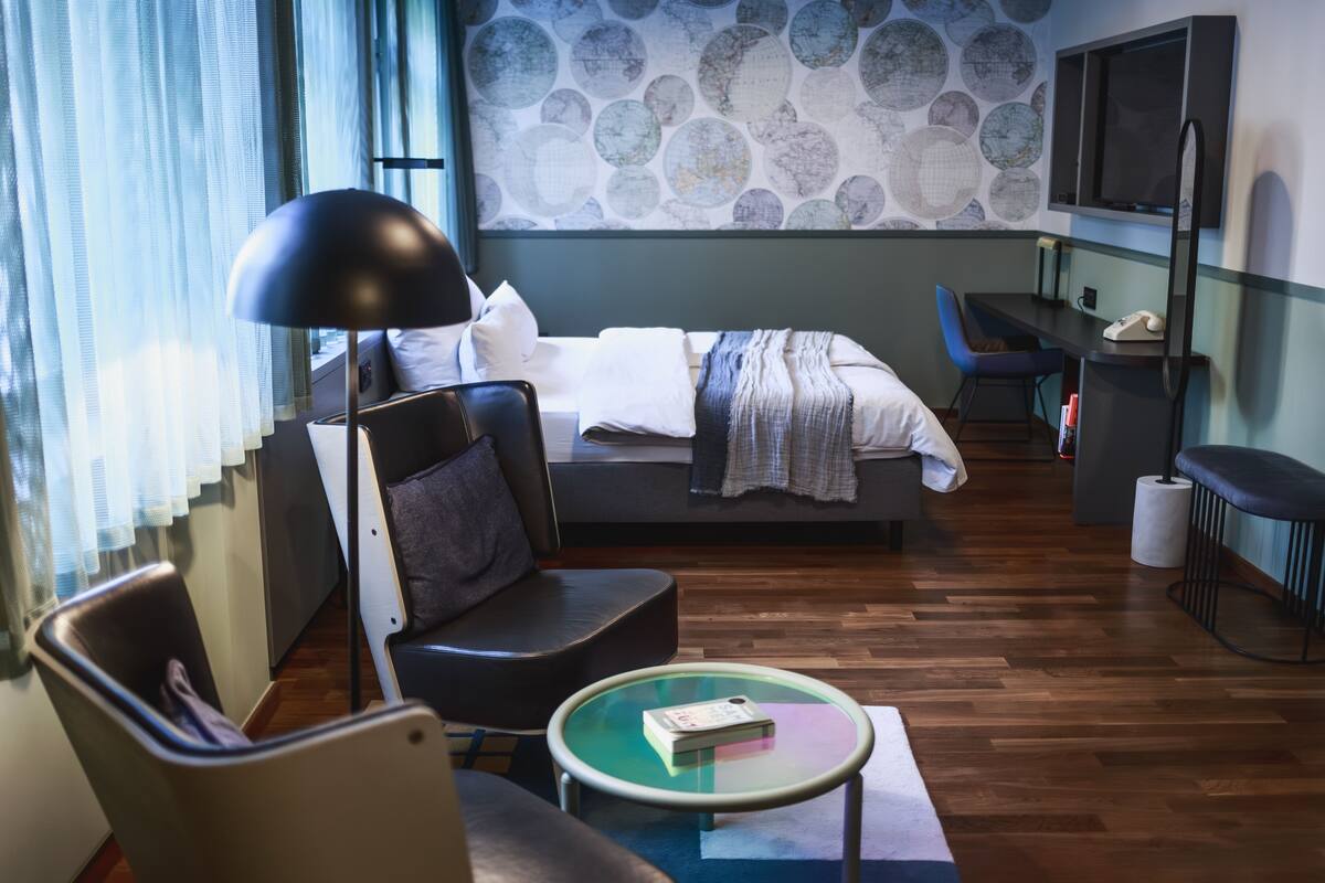 Ein modernes Hotelzimmer mit Bett, Schreibtisch, Sessel, rundem Tisch und einem großen Fenster mit Vorhängen.