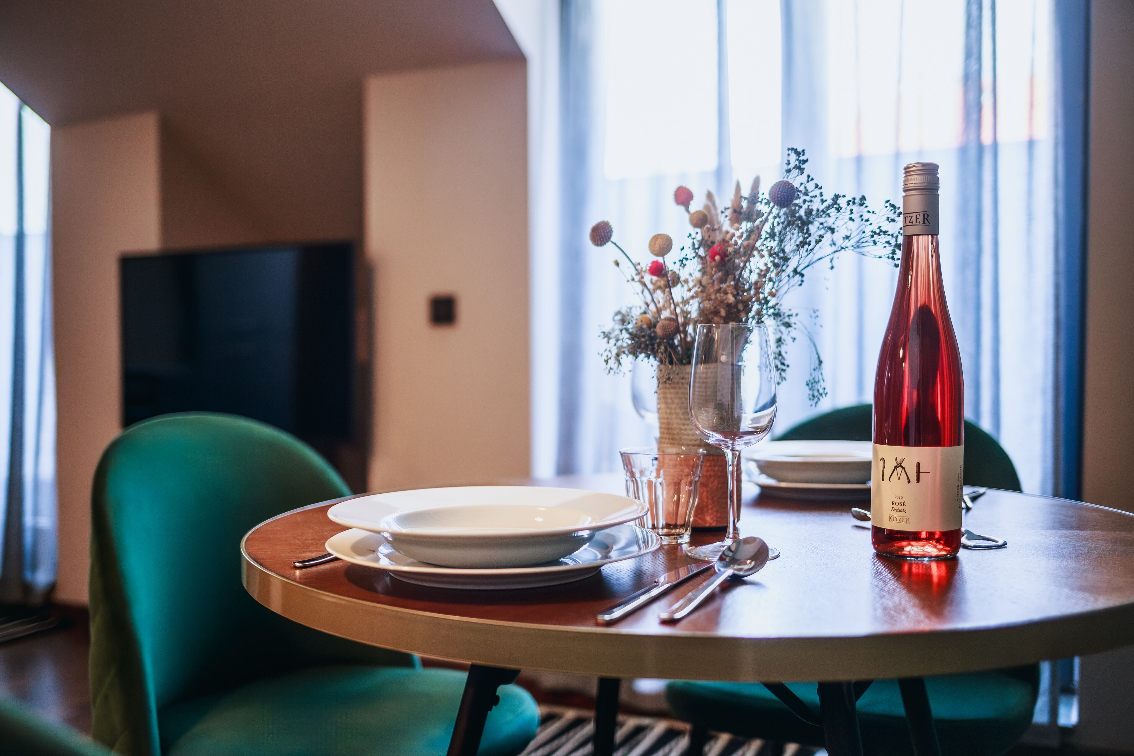 Una mesa de comedor con platos, cubiertos, una botella de vino y un florero con flores.
