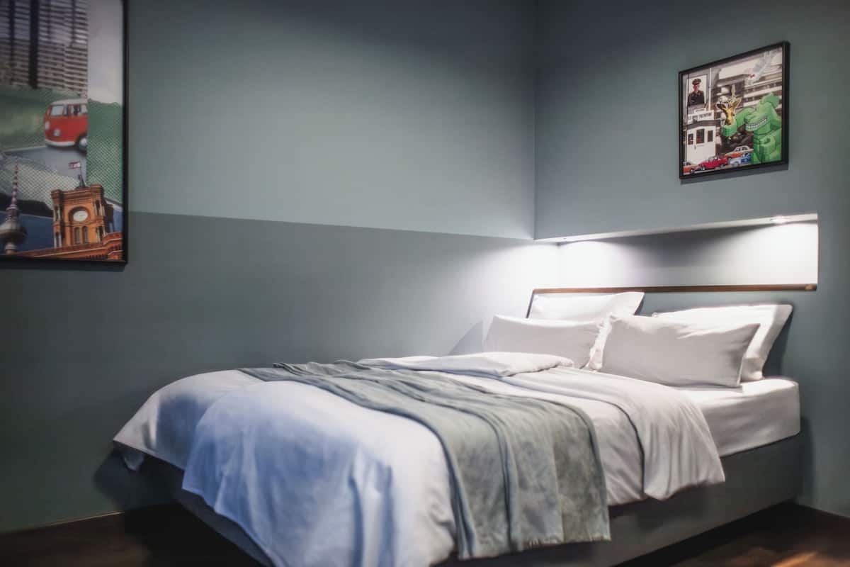 Ein ordentlich bezogenes Bett mit weißen Leinen und einer grauen Decke, eine an der Wand befestigte Lampe darüber und zwei gerahmte Bilder an der Wand.