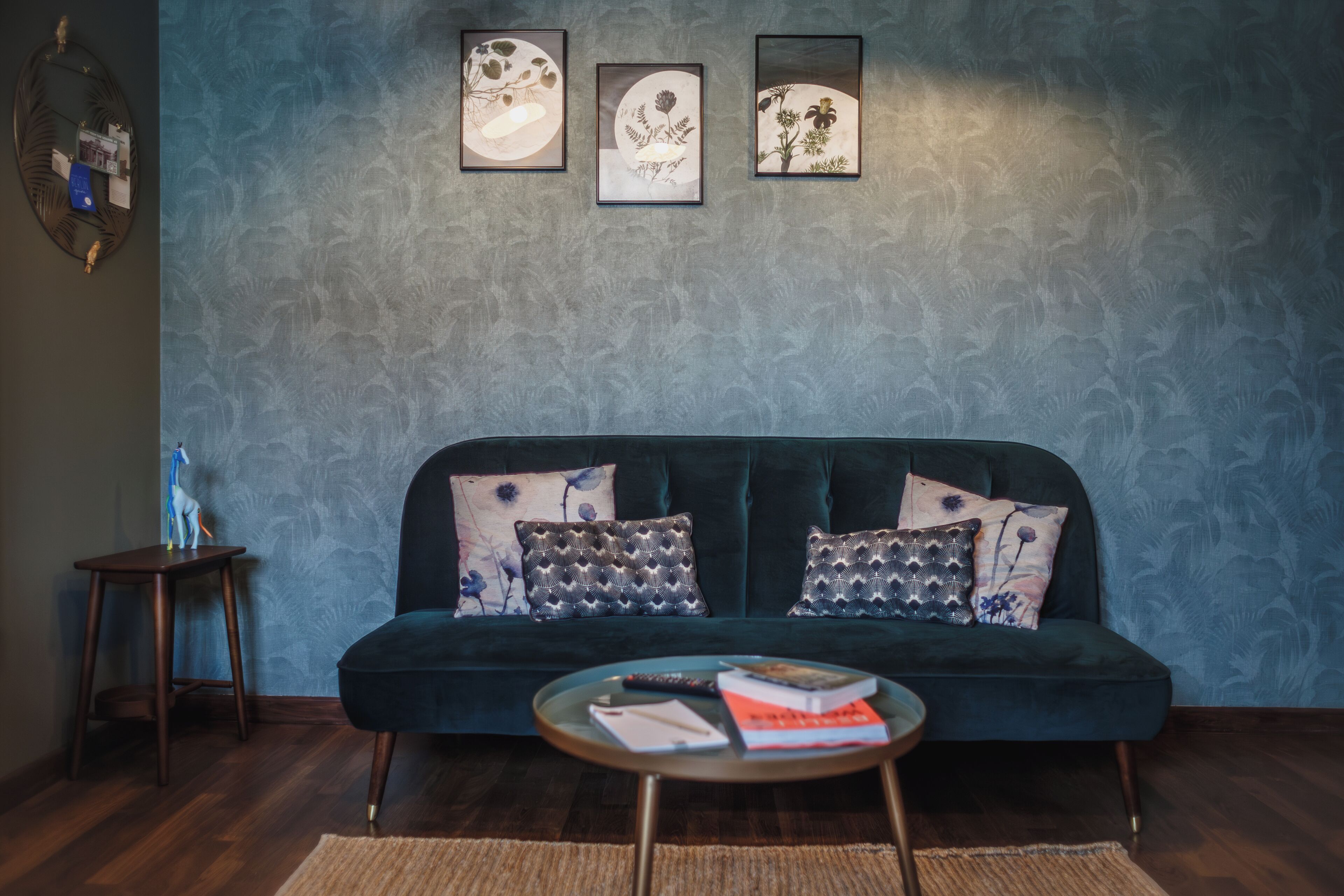Un divano verde scuro con cuscini decorati, un tavolino rotondo con libri e un telecomando, e tre quadri appesi al muro.