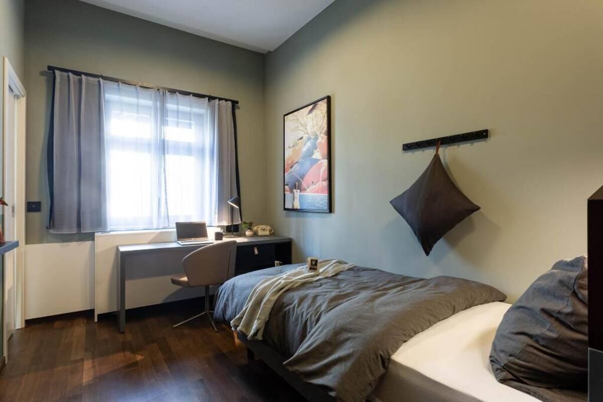 Ein Schlafzimmer mit Bett, Schreibtisch, Stuhl, einem Fenster mit Vorhängen und einem an der Wand befestigten Regal.
