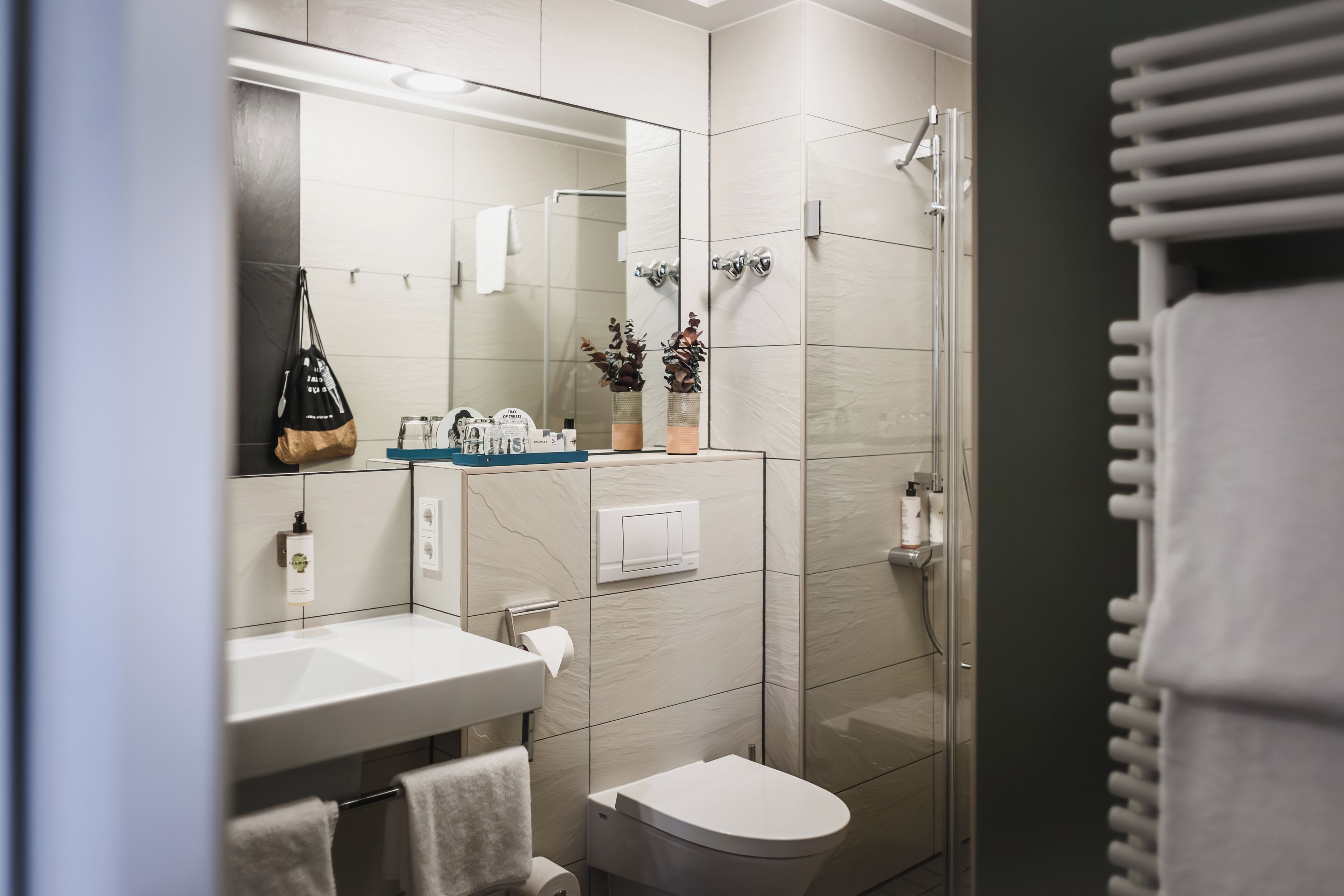 Un baño moderno con lavamanos, espejo y retrete blancos. Hay artículos de aseo en el lavamanos y un perchero para toallas.