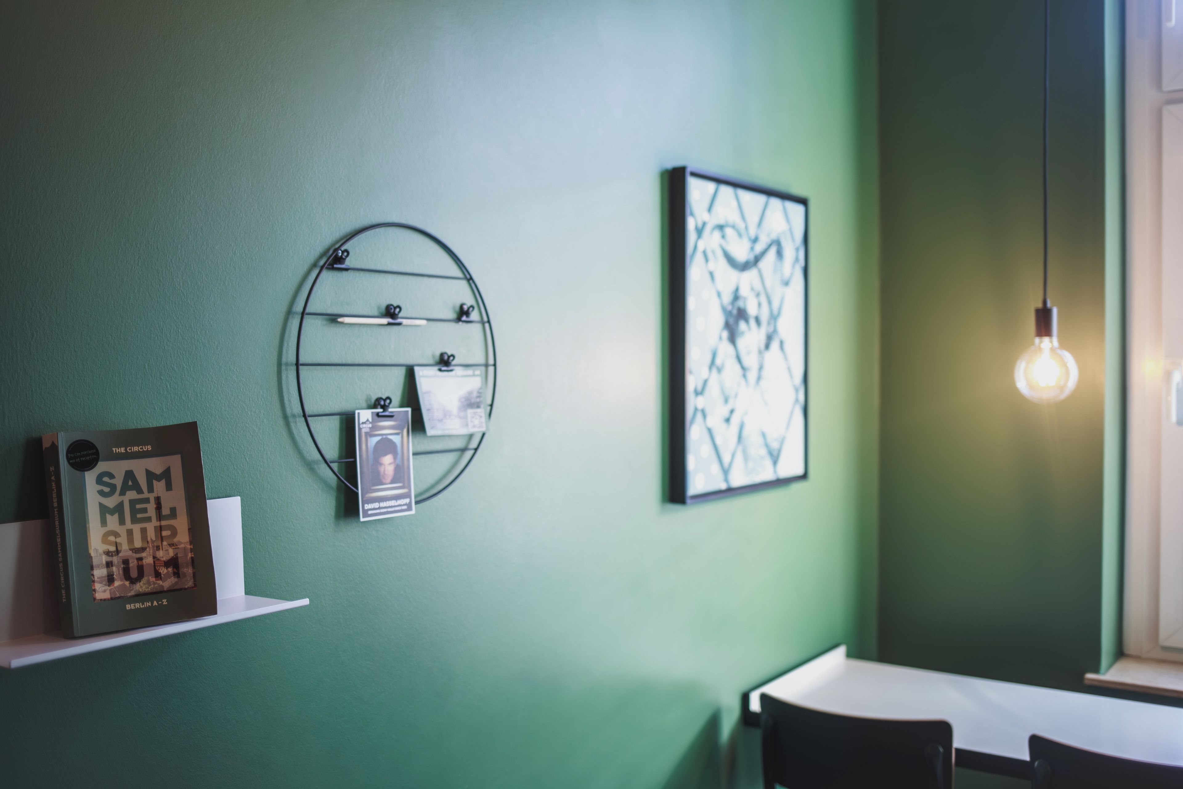 Una habitación con pared verde, estante metálico redondo con objetos, un cuadro enmarcado y una bombilla colgante.