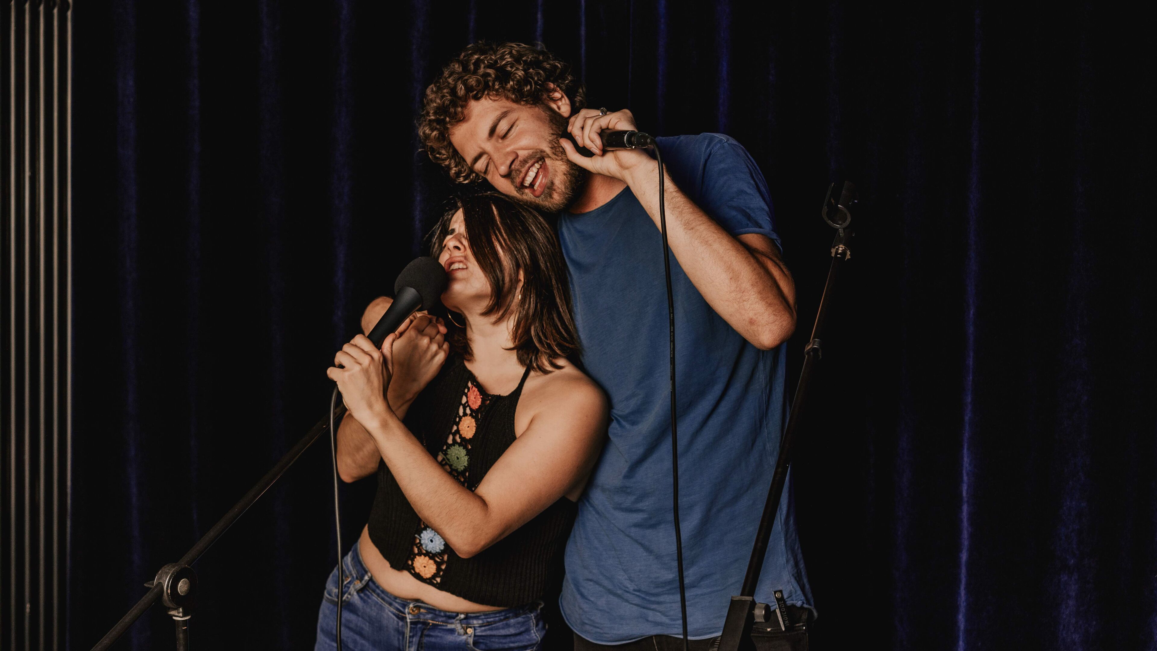 A person holding a microphone, another person standing behind them, and a guitar leaning against the wall.