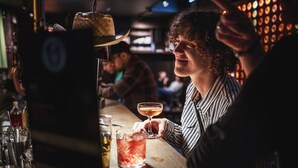 Eine Bar mit einem Barkeeper, der ein Getränk zubereitet, einem Hut auf der Theke sowie verschiedenen Flaschen und Gläsern.