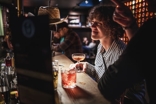 Uma área de bar com um bartender preparando uma bebida, um chapéu no balcão e diversos frascos e copos.