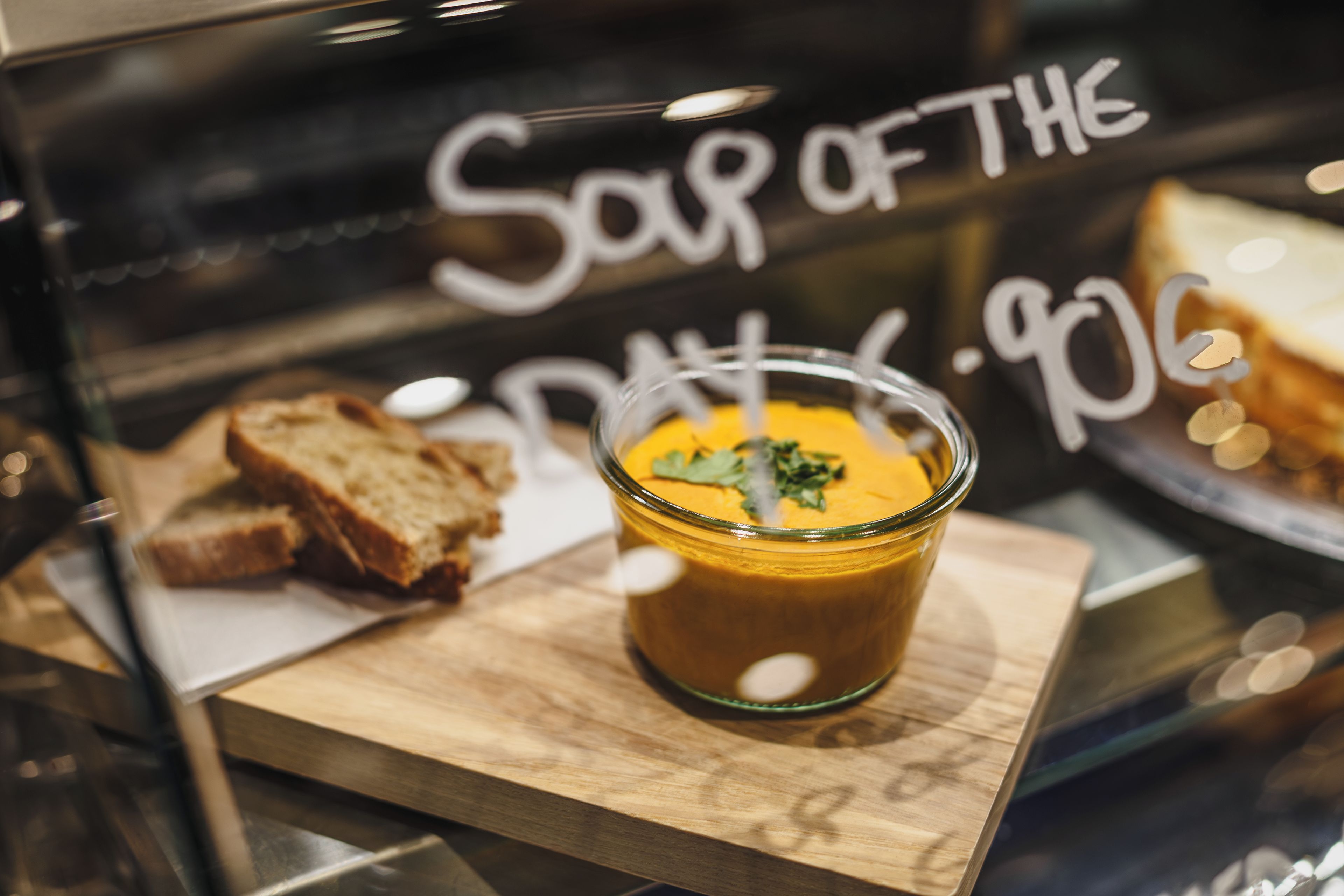 A glass of soup with a garnish on a wooden board, with bread and a sign in the background.