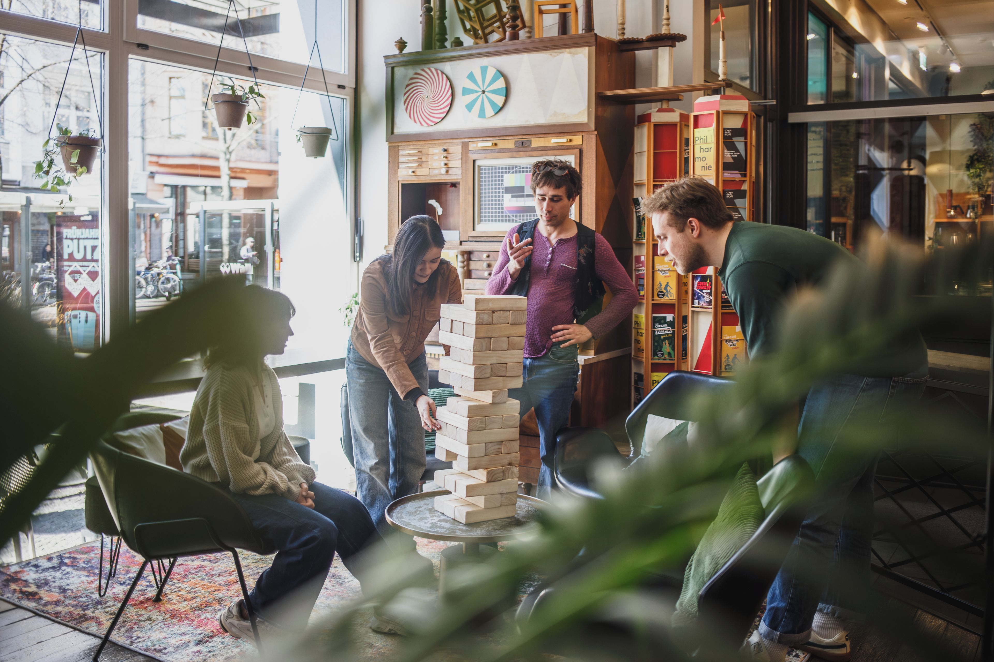 Un grupo de personas juega al Jenga en un espacio interior bien iluminado con amplios ventanales y una variedad de estanterías de libros al fondo.