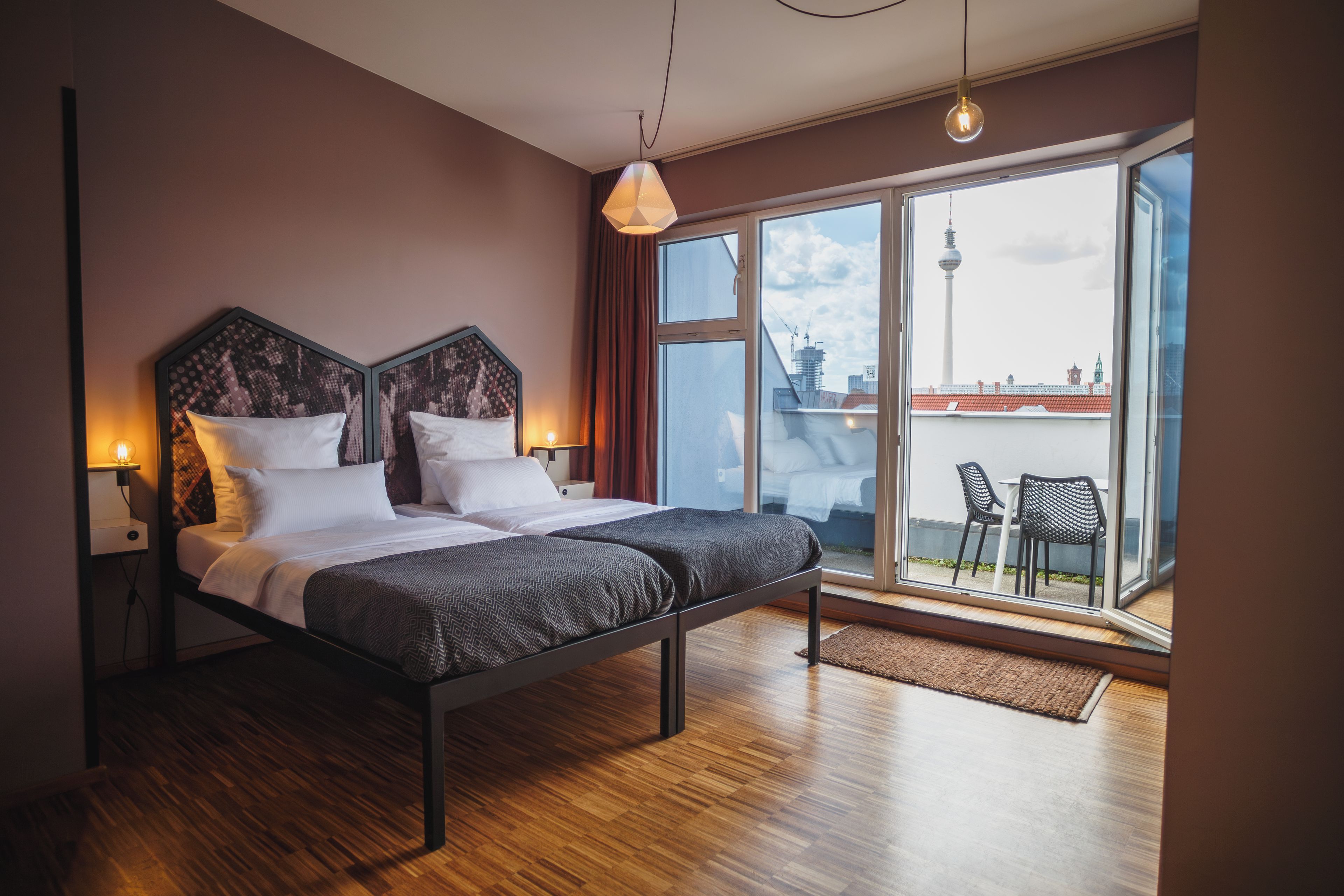 A modern bedroom with a large bed, a view of the city through a sliding door, and a balcony with outdoor furniture.