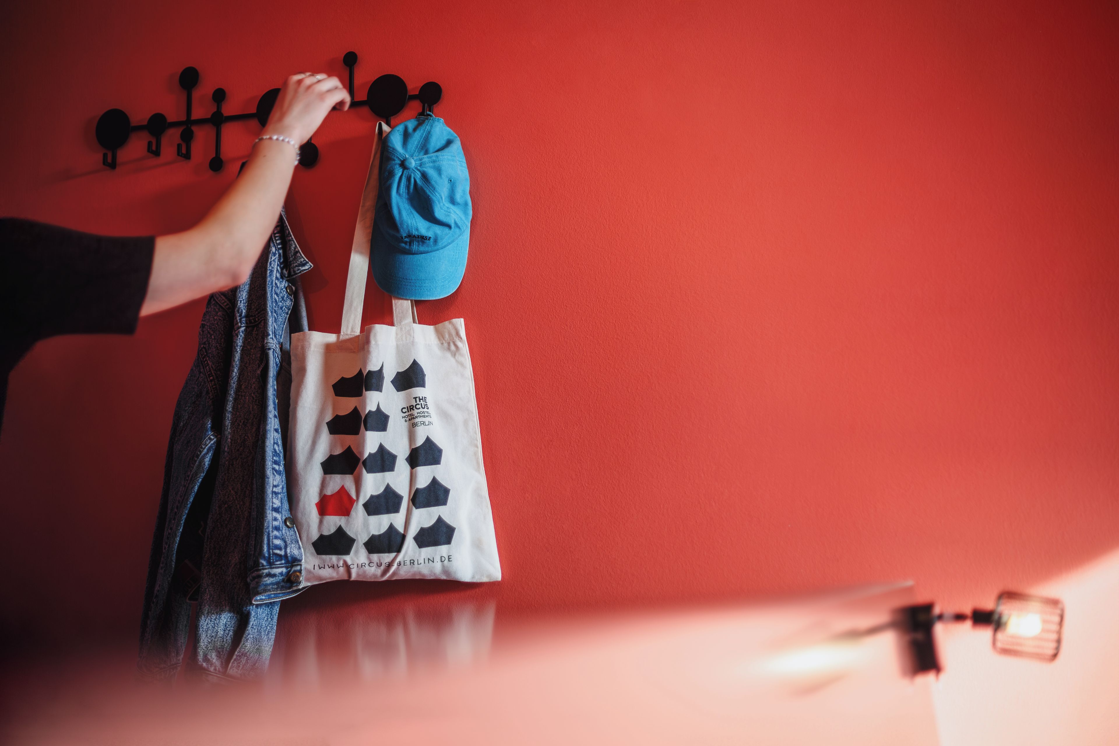 Una mano sosteniendo una bolsa azul y una bolsa de tela blanca con un estampado, frente a una pared roja con un perchero negro.