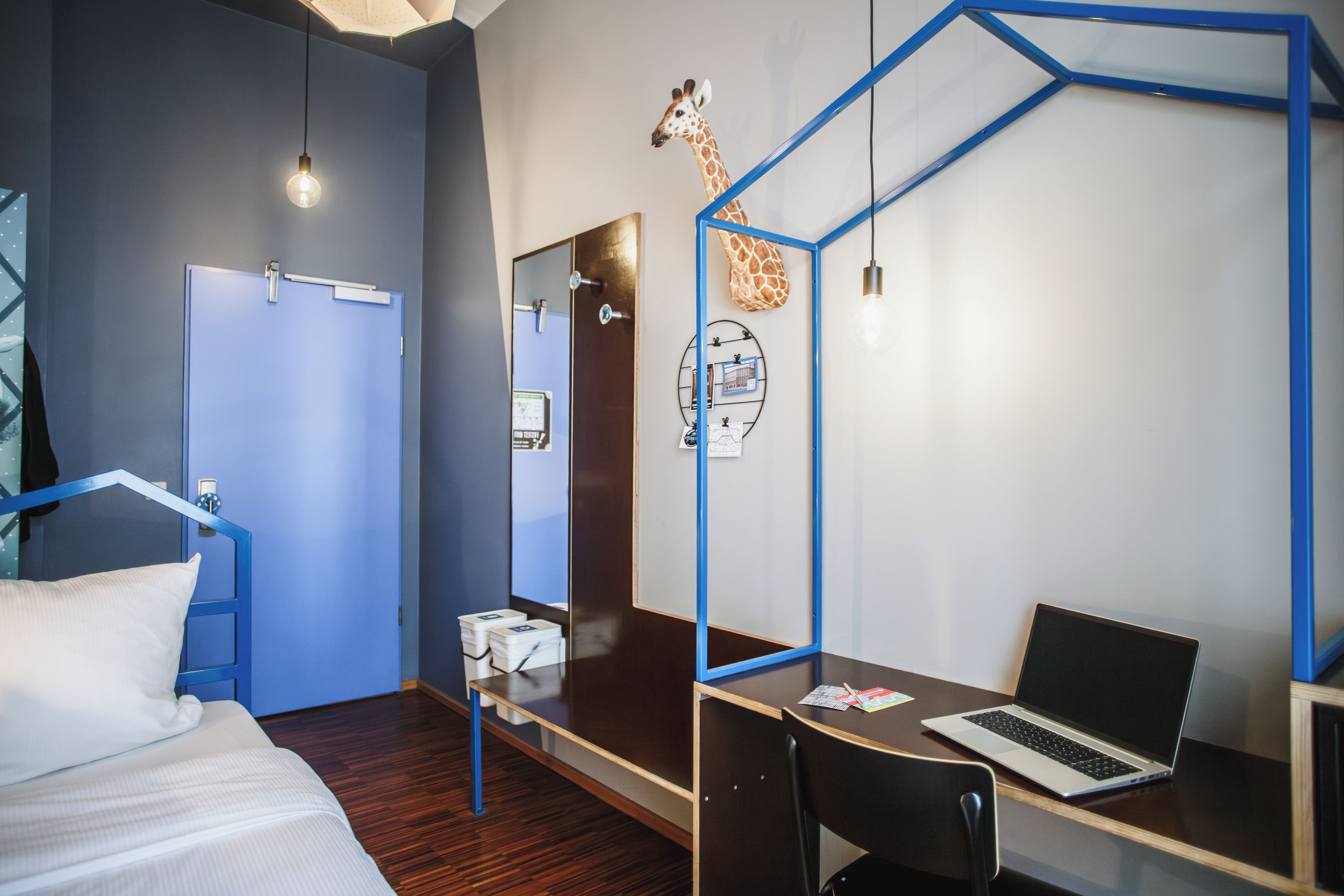 Une chambre d’hôtel comprenant un lit, un bureau avec un ordinateur portable, une chaise, une lampe de bureau, une sculpture de girafe fixée au mur et un miroir.