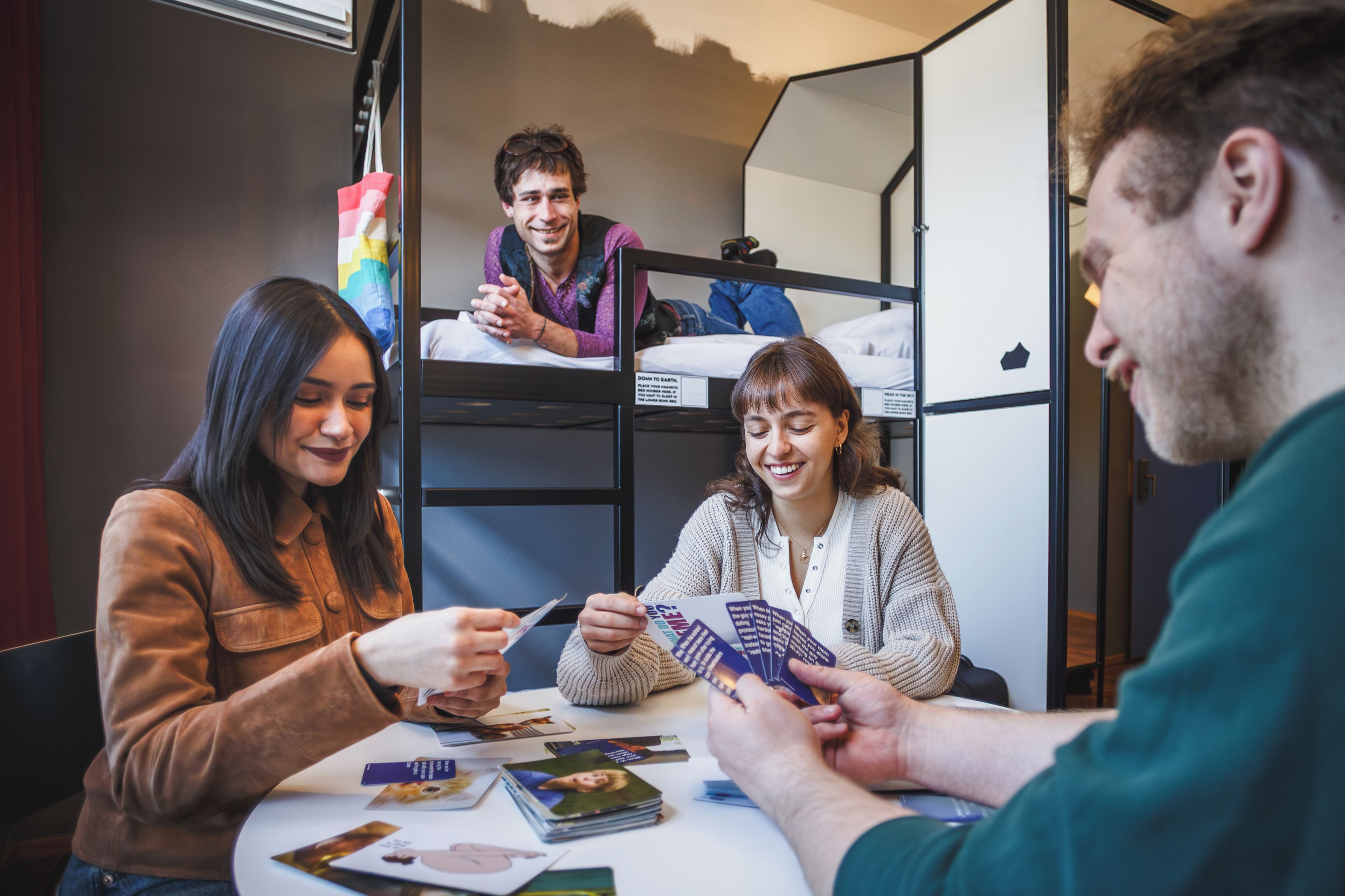 Un grupo de personas sentadas en una mesa con libros y cartas, en una habitación con literas y una ventana.