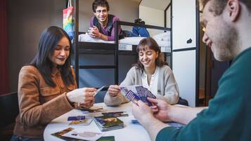 Un grupo de personas sentadas en una mesa con libros y cartas, en una habitación con literas y una ventana.