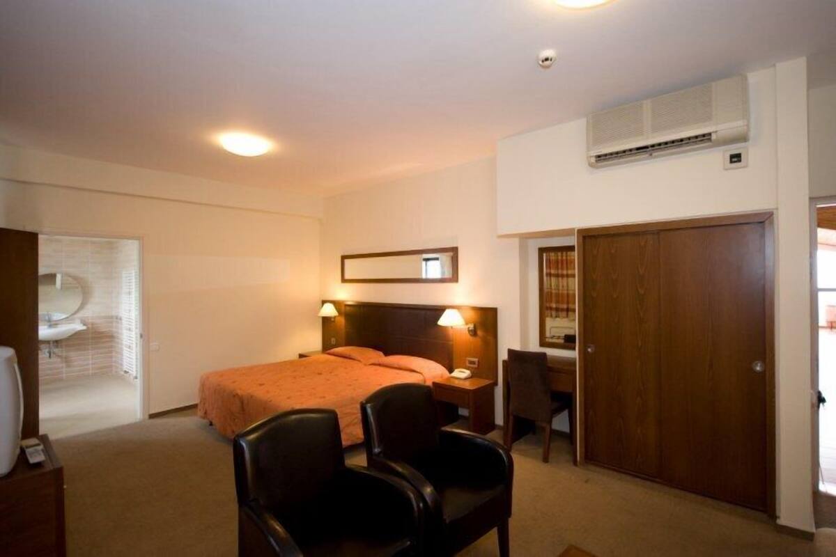 Ein Hotelzimmer mit einem Bett, zwei Nachttischlampen, einem Schreibtisch mit Stuhl, einem Kleiderschrank und einer Klimaanlage.