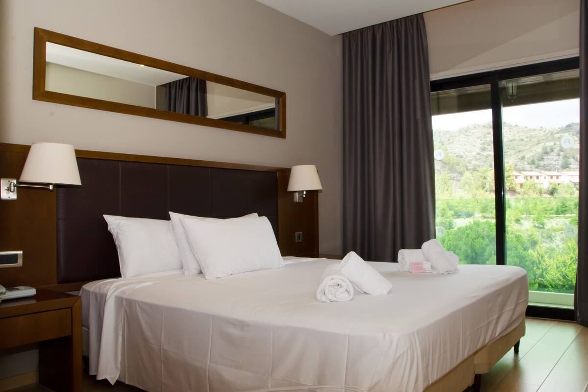 Ein Hotelzimmer mit einem großen Bett, zwei Nachttischlampen, einem Nachttisch mit Telefon, einem Spiegel über dem Bett und Blick auf Grün durch das Fenster.
