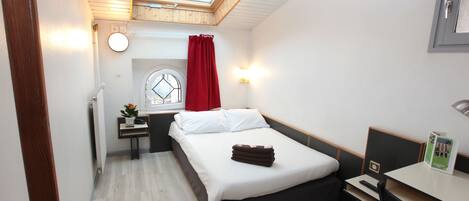 Una habitación de hotel compacta con tragaluz, una cama, un escritorio y una pequeña mesa con lámpara.