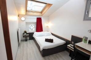A compact hotel room with a skylight, a bed, a desk, and a small table with a lamp.
