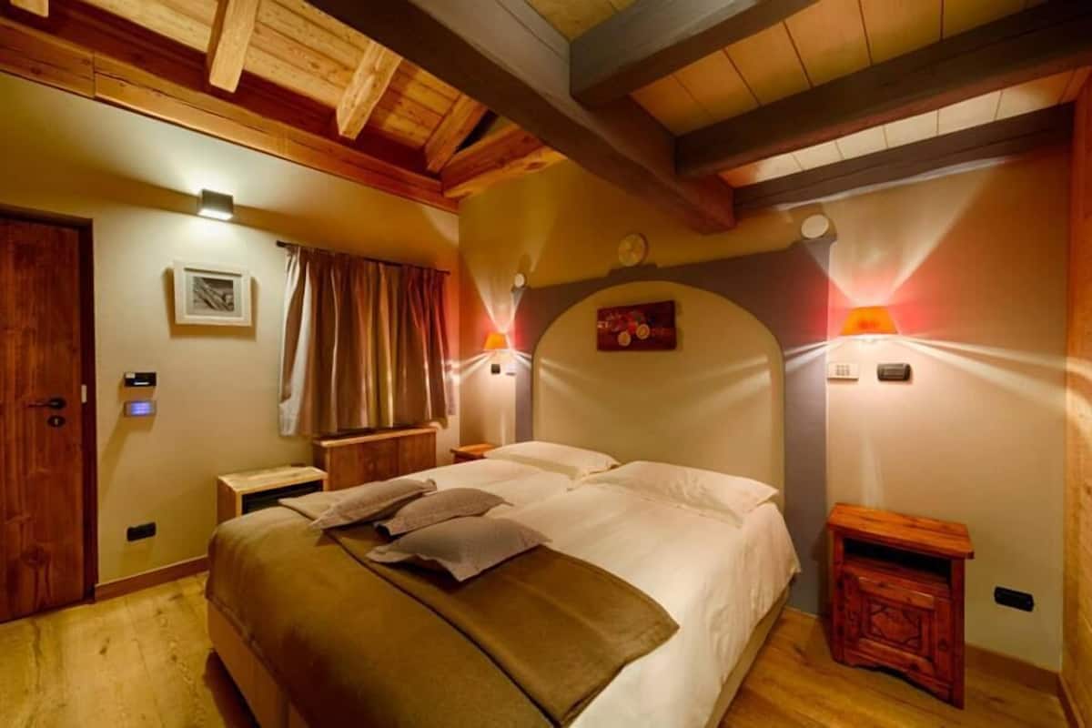 Un dormitorio acogedor con una cama grande, vigas de techo de madera, una mesita de noche, una lámpara de pared y un pequeño armario de madera.